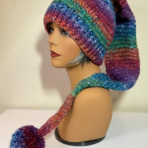 May include: A colorful, handmade crochet hat with a long, pointed end and a matching scarf with a pom-pom. The hat and scarf are a mix of rainbow colors, including blue, purple, green, and red. The hat is displayed on a mannequin head.