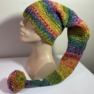 May include: A colorful, handmade crochet hat with a long, pointed tail and a pom-pom. The hat features a rainbow of colors, including yellow, orange, green, blue, and purple. The hat is displayed on a mannequin head.
