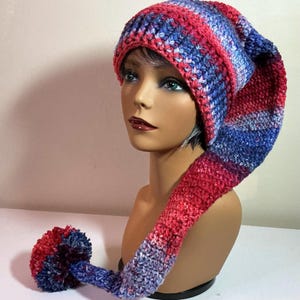 May include: A long, slouchy crochet hat featuring a variegated yarn pattern in shades of red, blue, and purple. The hat has a ribbed brim and a long, tapered tail that ends in a large, round pom-pom made from the same multi-colored yarn.