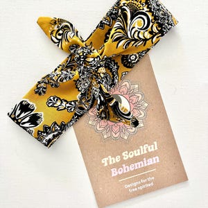 May include: Mustard yellow headband with a black and white floral design. The headband is knotted. A brown card with the text "The Soulful Bohemian" is next to the headband.