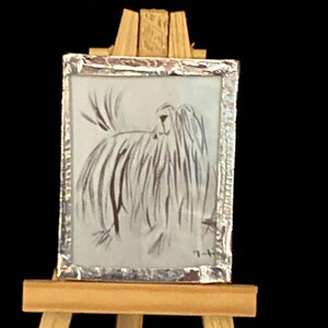 May include: A small framed black and white ink drawing of a dog on a wooden easel. The drawing is in a silver frame.
