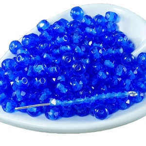 May include: A close-up of a pile of small, faceted, cobalt blue glass beads. The beads are arranged in a loose pile on a white surface.
