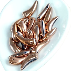 May include: A pile of rose gold colored glass beads in the shape of a teardrop with a pointed tip. The beads are arranged on a white heart-shaped dish.