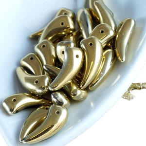 May include: A pile of gold-colored, shiny, plastic beads shaped like a curved fang or tooth. The beads are arranged in a bowl.