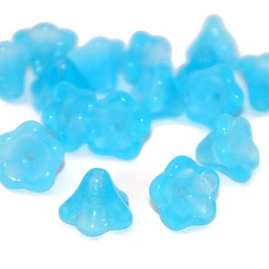 May include: A group of 14 light blue glass flower-shaped beads. The beads have a frosted finish and are scattered on a white surface.