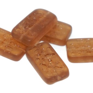 Könnte beinhalten: Fünf braune, durchsichtige Bonbons in Form von rechteckigen Stäben mit erhabenen Buchstaben. Die Bonbons liegen auf einem weißen Untergrund.