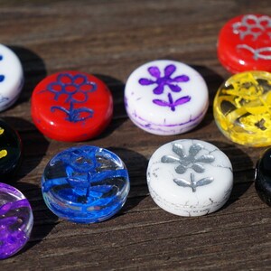 May include: Ten round glass tiles with different coloured backgrounds and flower designs. The designs are painted in white, yellow, blue, purple, silver, and black.