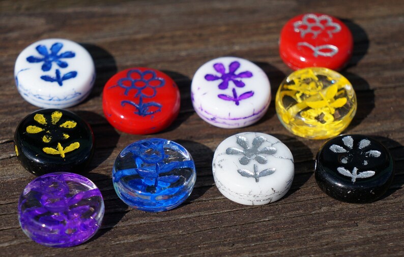 May include: Ten round glass tiles with different coloured backgrounds and flower designs. The designs are painted in white, yellow, blue, purple, silver, and black.