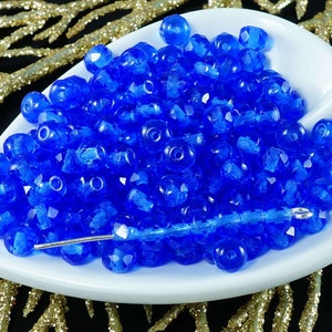 May include: A close-up of a pile of small, faceted, blue glass beads. The beads are arranged in a white bowl and are illuminated by a bright light.