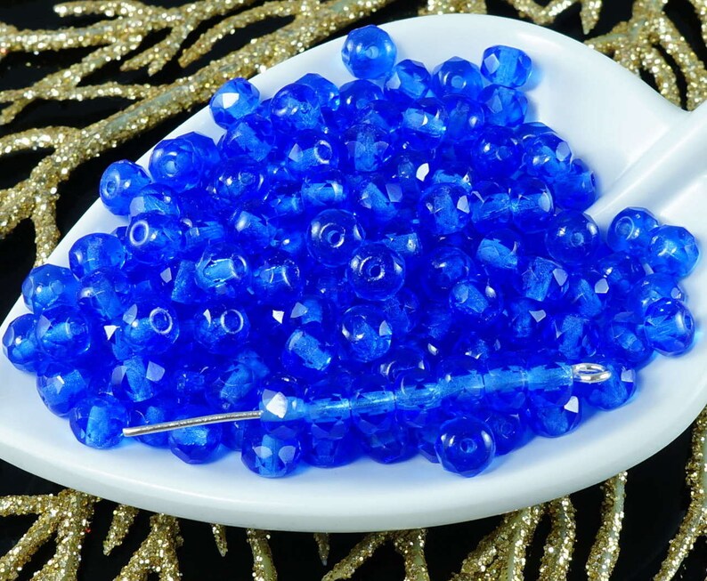May include: A close-up of a pile of small, faceted, blue glass beads. The beads are arranged in a white bowl and are illuminated by a bright light.