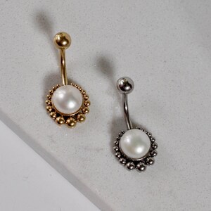 Può includere: Due piercing all'ombelico. Uno è dorato con una perla e piccole perline dorate. L'altro è argentato con una perla e piccole perline argentate. Entrambi hanno un design curvo.