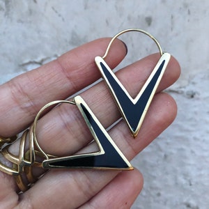 May include: A pair of gold-toned hoop earrings featuring a V-shaped design with a smooth black inlay. The earrings have a thin, circular wire post that connects to the top of the triangular frame, creating a geometric look with clean lines.