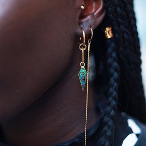Peut inclure: Deux créoles dorées portées au lobe de l'oreille. L'une des créoles présente une breloque dorée suspendue avec une incrustation de nacre d'ormeau irisée, verte et bleue, en forme de losange. L'autre créole est reliée à une longue chaîne dorée.