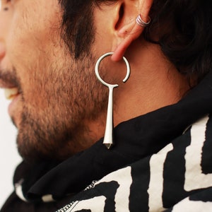 May include: A silver-toned earring featuring a circular hoop top that transitions into a smooth, elongated, tapered drop pendant. The earring is worn in the earlobe, while a separate, smaller silver-toned triple-band cuff earring is worn in the upper ear cartilage.