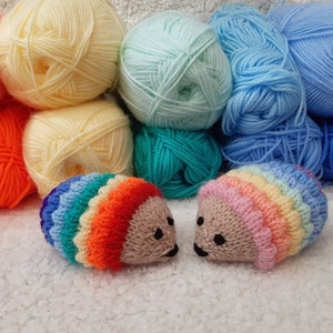 May include: Two rainbow coloured knitted hedgehogs with black eyes and smiling mouths. The hedgehogs are sitting on a white fluffy surface. There are balls of yarn in the background in various colours, including orange, yellow, blue, green, and pink.