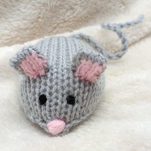 May include: A knitted mouse toy in grey, pink, and black. The mouse has large pink-lined ears, small black eyes, and a pink nose. A grey knitted tail extends from the back. The toy is set against a soft, white background.