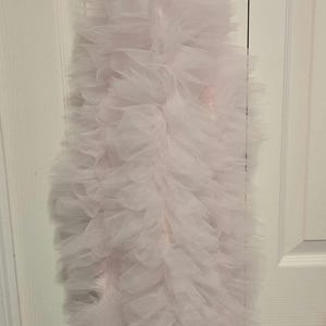 May include: A light pink, heart-shaped decoration hangs from a white hook on a white door. The decoration is made of a fluffy, layered, light pink material, resembling a scarf or boa.