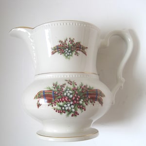 May include: White ceramic pitcher with a gold trim and a Christmas-themed design featuring red berries, green leaves, and a plaid ribbon.