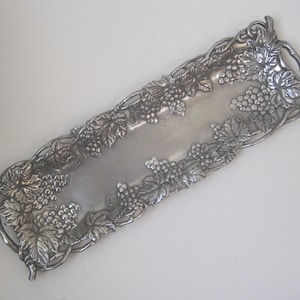 May include: A rectangular silver-toned serving tray with a grape and leaf design around the edges. The tray has a smooth, reflective surface in the center and handles on each end. The grape clusters and leaves are intricately detailed.