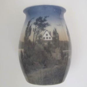 May include: A ceramic vase with a landscape scene. The vase has a blue gradient at the top, transitioning to a scene of trees, a house, and water. The artwork is in shades of gray and brown.