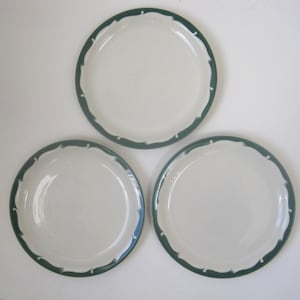 May include: Three white ceramic plates with a dark teal scalloped border. The plates are round and have a simple, elegant design. The plates are arranged in a triangular pattern.