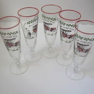 May include: Five clear champagne flutes with red rims. Each glass features a vintage car design with the text "Buick model 6 1908" and decorative green leaves. The glasses are on a white surface.