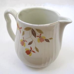 May include: A white ceramic creamer with a floral pattern in shades of brown, orange, and yellow. The creamer has a rounded body and a curved handle.