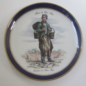May include: A white ceramic plate with a blue and gold rim. The plate features a colorful illustration of a man carrying two buckets and the text "Metiers du Vieux Paris" and "Marchand de Sauces Fraises".
