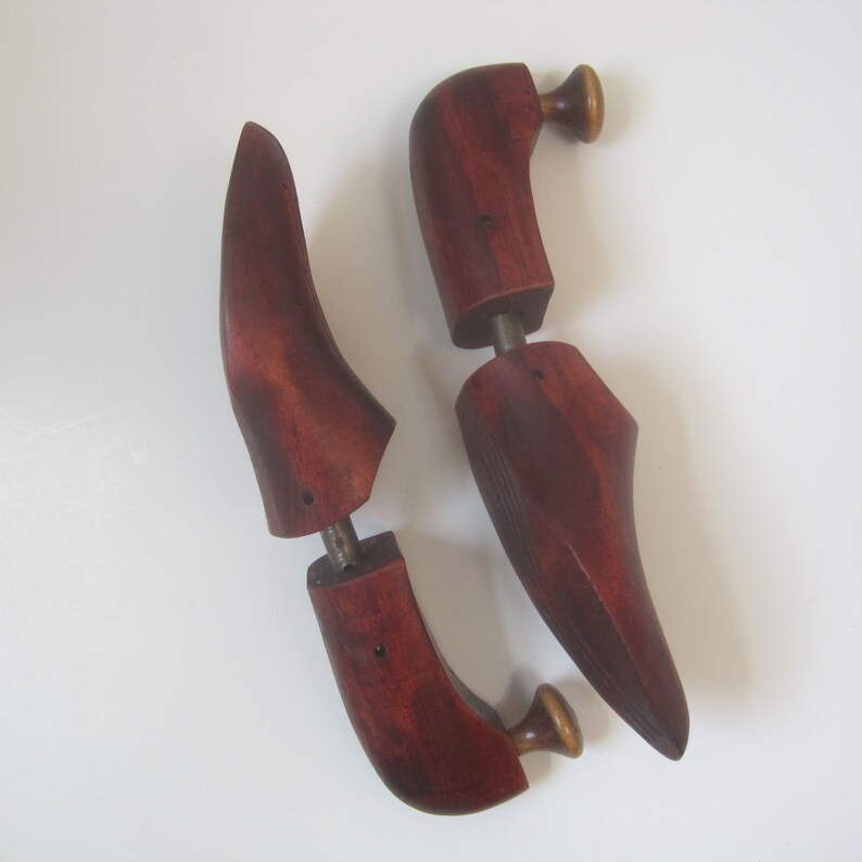 Puede incluir: Un par de hormas de zapatos de madera con un acabado rojo oscuro. Las hormas tienen un pomo en la parte superior y una varilla de metal que conecta las dos piezas.