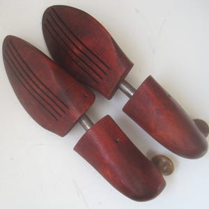 K&ouml;nnte beinhalten: Ein Paar Schuhspanner aus Holz mit einer rotbraunen Oberfl&auml;che. Die Schuhspanner haben eine Metallstange, die die beiden H&auml;lften verbindet, und einen Knopf an einem Ende.