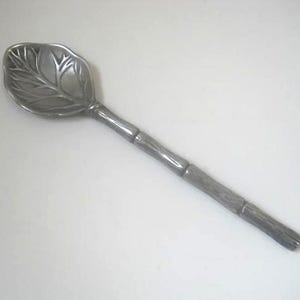 May include: A silver-colored serving spoon with a leaf-shaped bowl and a bamboo-style handle. The leaf bowl features detailed veining. The handle has a textured, segmented design. The spoon is made of metal.
