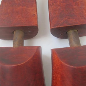 K&ouml;nnte beinhalten: Ein Paar Holzschuhspanner mit einer rotbraunen Oberfl&auml;che. Die Schuhspanner haben eine abgerundete Oberseite und einen Metallstab in der Mitte.