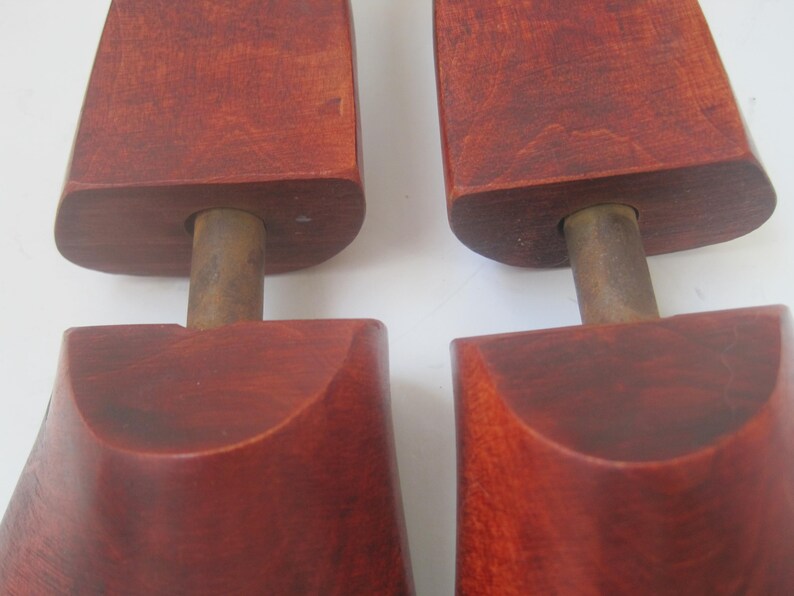 K&ouml;nnte beinhalten: Ein Paar Holzschuhspanner mit einer rotbraunen Oberfl&auml;che. Die Schuhspanner haben eine abgerundete Oberseite und einen Metallstab in der Mitte.