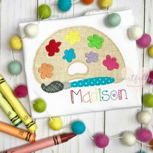 May include: White t-shirt featuring an embroidered artist's palette with colorful paint splotches and a paintbrush. The name "Madison" is stitched in rainbow thread below the palette. Decorative felt ball garland and crayons surround the shirt.