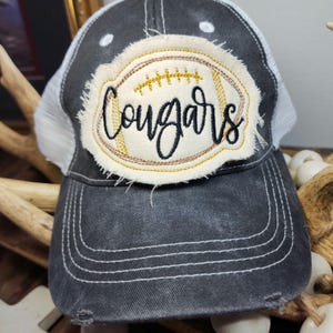 Pode incluir: Um boné de beisebol cinza desgastado com uma parte traseira de malha branca. A frente apresenta um patch bordado em forma de bola de futebol com a palavra "Cougars" em escrita preta. O patch tem detalhes dourados e creme.
