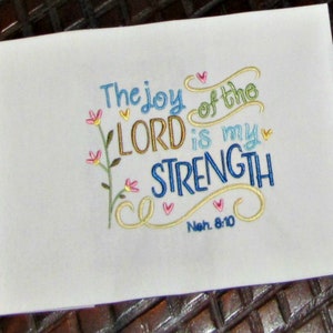 May include: Embroidered white fabric with the text "The joy of the LORD is my STRENGTH" in colorful cursive lettering. The text is surrounded by decorative swirls and hearts. The verse reference "Neh. 8:10" is included below the text.