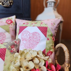 May include: Two pink and green patchwork throw pillows with a white embroidered paisley heart design. The pillows are sitting in a wicker basket with white hydrangeas.