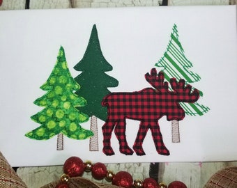 Könnte beinhalten: Ein roter und schwarzer karierter Elch-Applikator steht vor drei grünen Weihnachtsbäumen. Die Bäume sind mit verschiedenen Mustern verziert, darunter Punkte und Streifen.