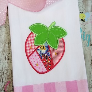 May include: A white kitchen towel with a pink and white gingham border. A red and green patchwork strawberry is embroidered on the towel.