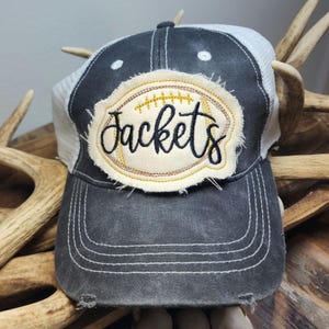 Pode incluir: Boné de caminhoneiro cinza e branco desgastado com um patch com um design de futebol americano e a palavra "Jackets" em escrita preta. O boné tem uma aba curva e uma parte traseira de malha.