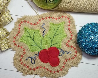 May include: A burlap Christmas ornament with a green holly leaf design and red berries. The ornament is decorated with red and blue stitching.