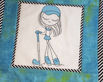 May include: A square quilt featuring an embroidered cartoon of a girl playing golf. The girl is wearing a blue visor, skirt, and shoes, and is holding a golf club. The quilt has a blue and green background with a black and white striped border.
