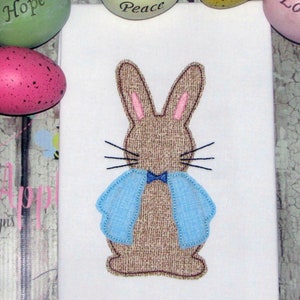 May include: Embroidered brown bunny with blue bow tie and light blue wings. The bunny is on a white background.