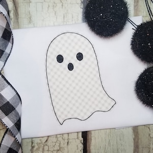 May include: A white ghost with black eyes and a mouth, embroidered on a white fabric. The ghost has a checkered pattern.