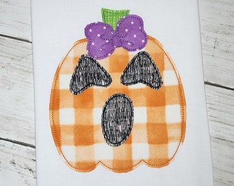 May include: A white fabric with an orange and white checkered pumpkin with a purple bow and a green stem. The pumpkin has a black stitched face with eyes, a mouth, and a nose.