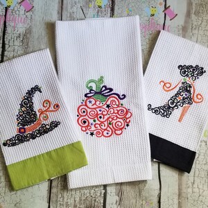 May include: Three white kitchen towels with embroidered Halloween designs. The designs are a witch's hat, a pumpkin, and a high-heeled shoe. Each design is made up of black, orange, green, and purple swirls.