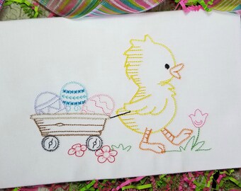 May include: White Easter-themed embroidered fabric featuring a yellow chick pulling a wooden wagon filled with decorated Easter eggs. The design includes flowers and grass, with colorful ribbon in the background.