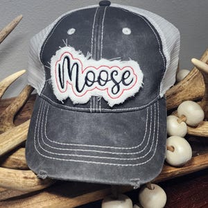 Pode incluir: Boné cinza de caminhoneiro com parte traseira em tela branca e aba desgastada. O boné apresenta a palavra "Moose" em um patch branco com borda vermelha. Costuras brancas acentuam o design.