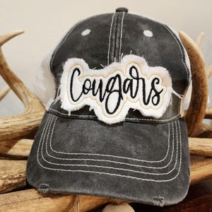Pode incluir: Um chapéu de caminhoneiro preto e branco com um remendo branco que diz "Cougars" em letras cursivas pretas. O remendo tem um contorno dourado.
