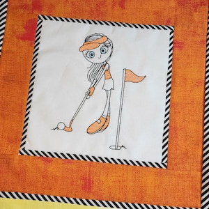 May include: Embroidered design of a girl playing golf. She is wearing a hat, a short-sleeved shirt, and a skirt. She is holding a golf club and is about to hit a golf ball. There is a golf flag in the background.